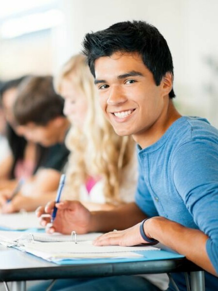 male student working on school work at desk in classroom and looking and smiling at camera